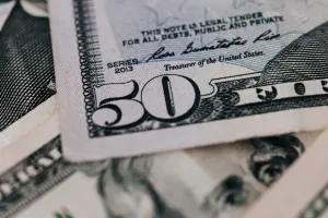 Dłoń trzymająca trzy banknoty dolarowe obok smartfona z prostym kalkulatorem walut na jasnym biurku.