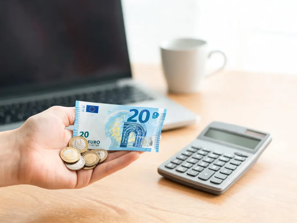 Dłoń trzymająca monety euro przy banknocie 20 € i kalkulatorze, symbolicznie ukazująca przeliczanie euro na złotówki.